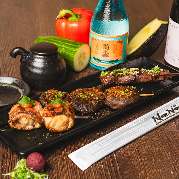 Japanese skewers, including scallops and beef, with sake bottles, vegetables, and chopsticks on a wooden table.