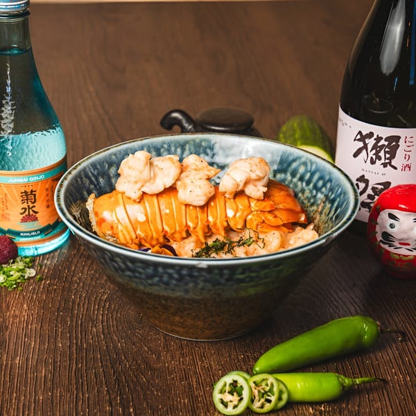 Lobster tail and shrimp in a bowl with two sake bottles, green peppers, and a Daruma doll.