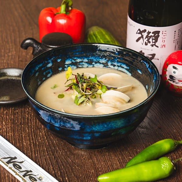 Clam chowder in a blue bowl, garnished with microgreens, alongside a sake bottle and peppers.