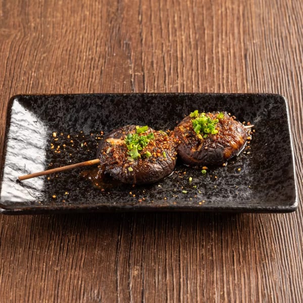 Two grilled mushrooms on a skewer, topped with green onions and spices, on a black rectangular plate.