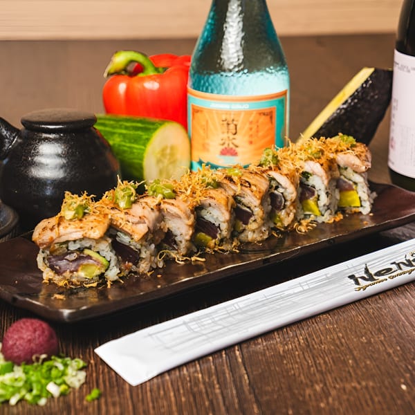Sushi rolls with seared fish, avocado, and crispy toppings, alongside vegetables and sake bottles.