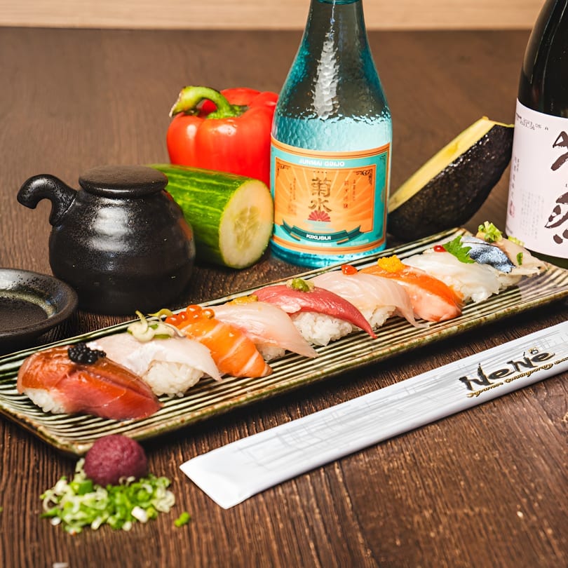A platter of colorful nigiri sushi, sake bottles, and fresh vegetables on a wooden table.
