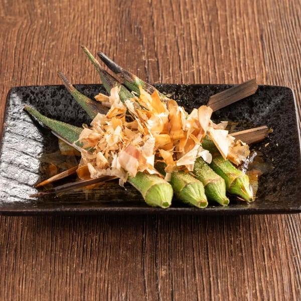 Grilled okra skewers topped with bonito flakes on a black rectangular plate.