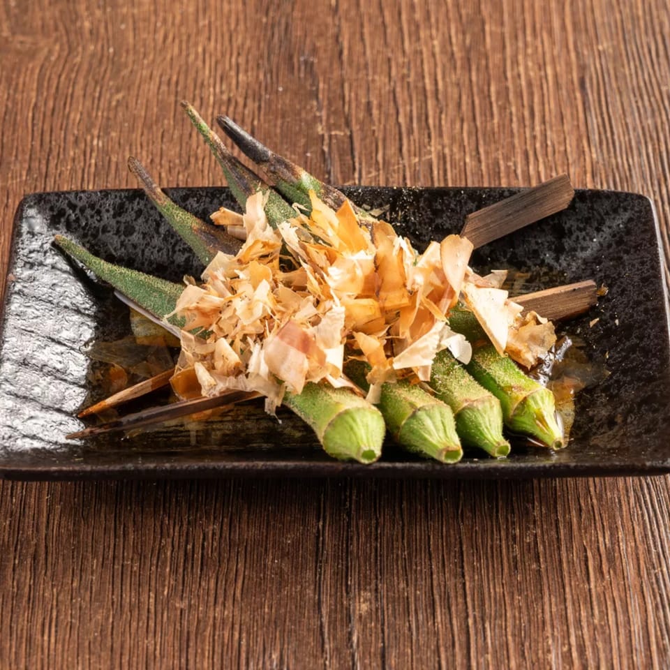 Grilled okra skewers topped with bonito flakes on a black rectangular plate.