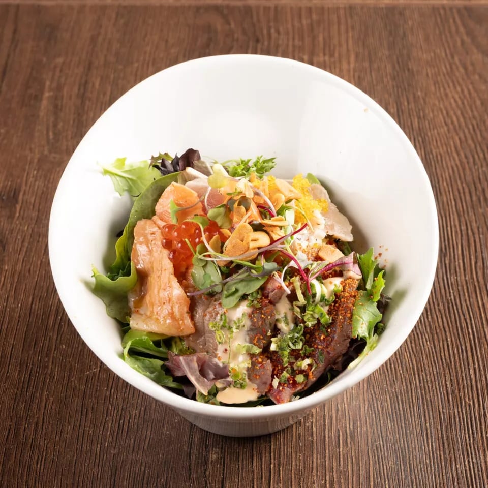 A white bowl filled with a colorful salad featuring greens, salmon, tuna, roe, and a savory dressing.