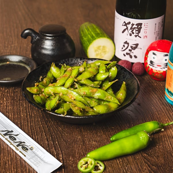 A bowl of edamame with a side of peppers, cucumber, and a bottle of sake.