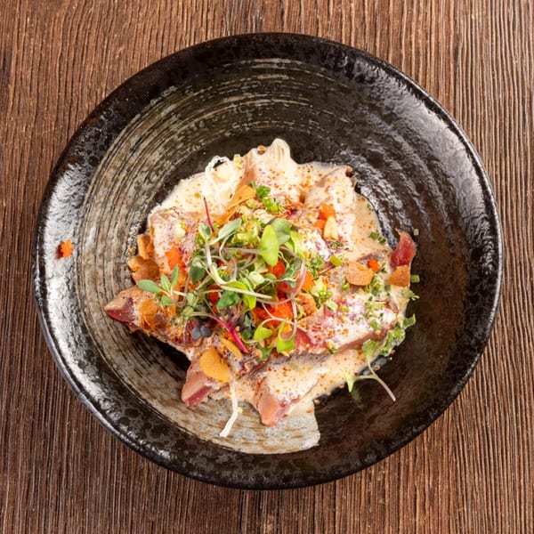 Overhead view of a dark bowl filled with sliced fish, creamy sauce, microgreens, and spices.
