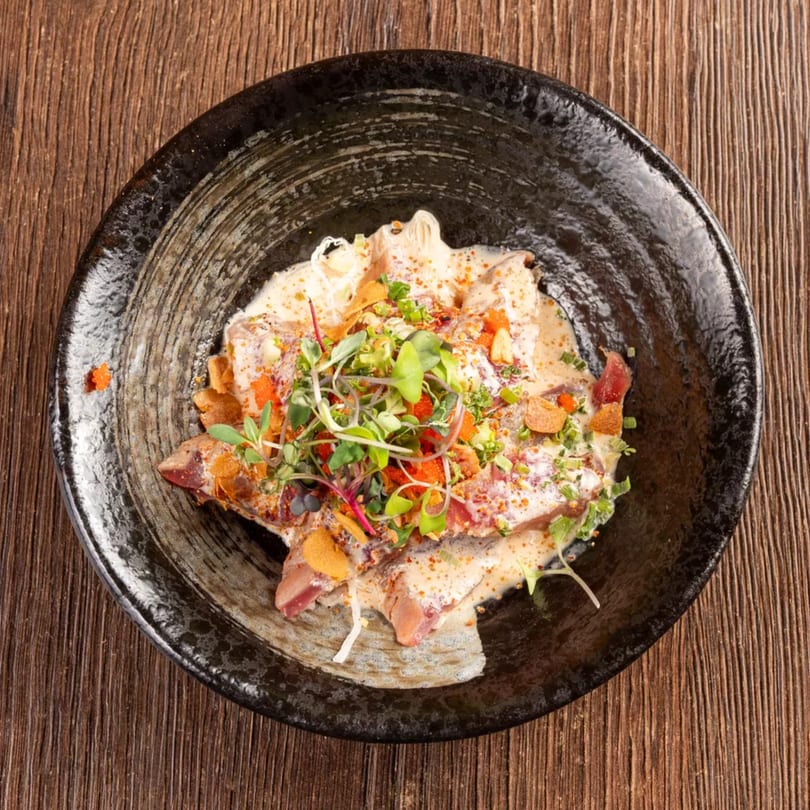 Overhead view of a dark bowl filled with sliced fish, creamy sauce, microgreens, and spices.
