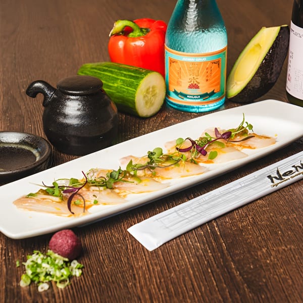 Sashimi with microgreens on a white plate, alongside sake, vegetables, and chopsticks on a wooden table.