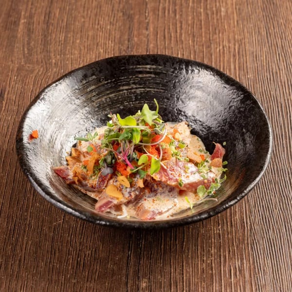 Seared tuna sashimi with creamy sauce, microgreens, and orange tobiko in a black bowl.